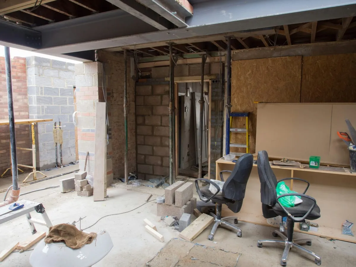 Two office chairs and a desk unit standing in a room under construction, among props, blockwork and timber offcuts.